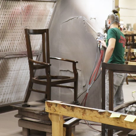 A catalyzed lacquer is being hand-sprayed onto the wooden chair frame